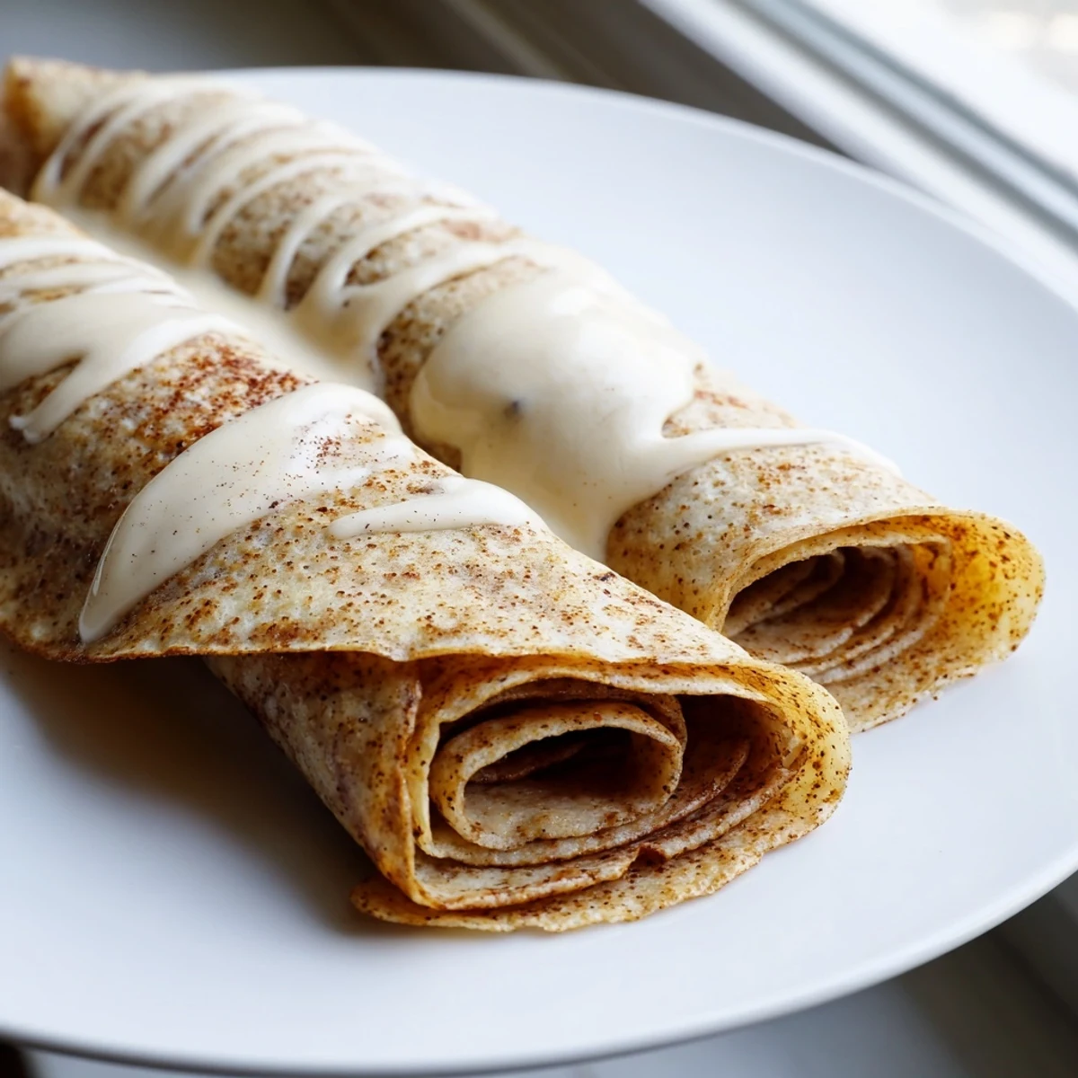 Rolled cinnamon roll protein crepes topped with tangy cream cheese glaze and a dusting of cinnamon